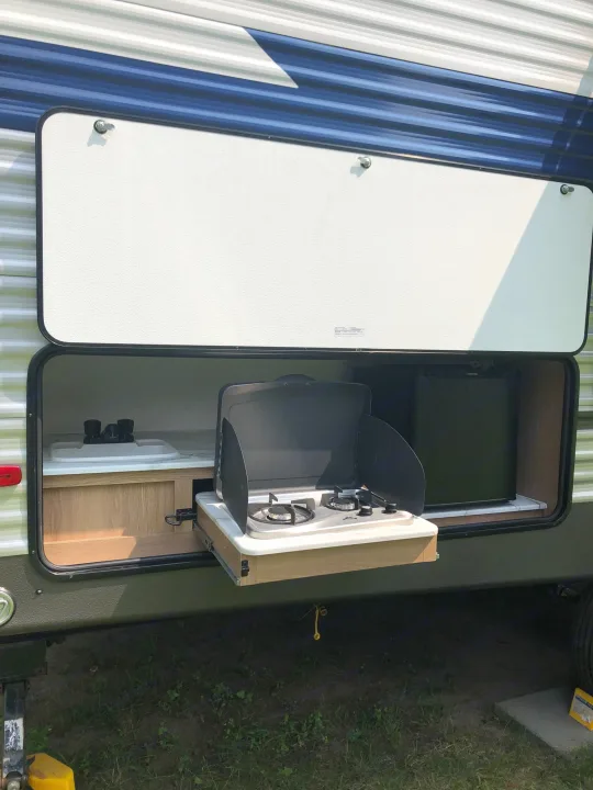 Side of an RV rental showing an open outdoor kitchen with a pull-out stove, sink, and compact fridge setup.. Forest River Prime Time Avenger 24BHS 2023