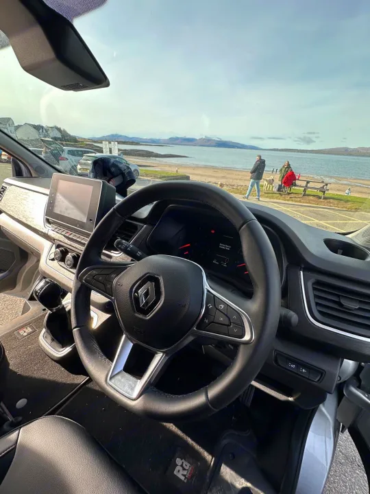 Inside a Renault RV rental, steering wheel and dashboard frame a scenic bay with mountains and people.. Renault Other 2023