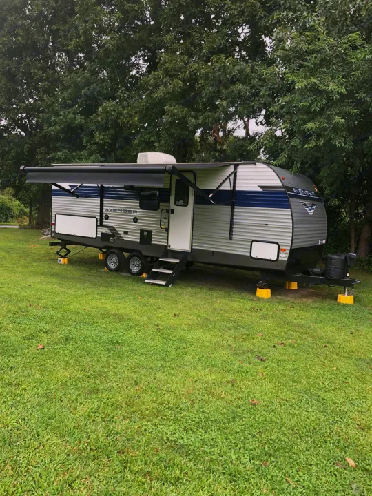 This gray and blue Avenger travel trailer, an ideal RV rental, is parked on lush green grass surrounded by trees for a perfect outdoor camping experience.. Forest River Prime Time Avenger 24BHS 2023