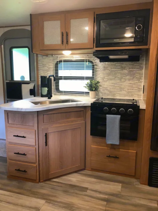 Step inside this modern RV rental kitchen, featuring light wood cabinets, a sink, microwave, and stove.. Forest River Prime Time Avenger 24BHS 2023