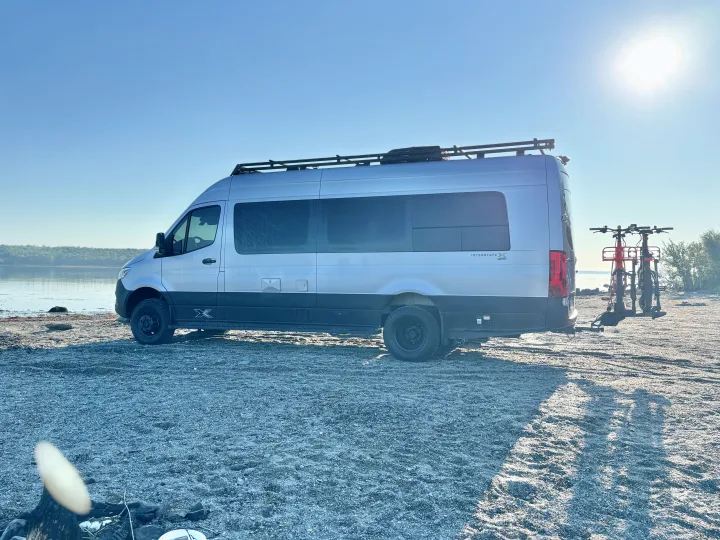 This silver Mercedes-Benz Sprinter rv rental camper van is parked on a gravel shore with bikes, perfect for outdoor adventures.. Airstream Interstate 2023