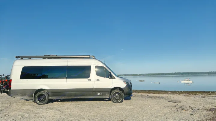 White RV rental van with bikes on a gravel beach. Peaceful bay scene with boats & distant treeline under blue sky.. Airstream Interstate 2023