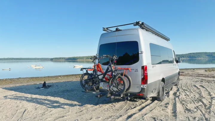 Silver Airstream RV rental camper van with bikes on a rack, backed onto a sandy beach by a tranquil bay.. Airstream Interstate 2023