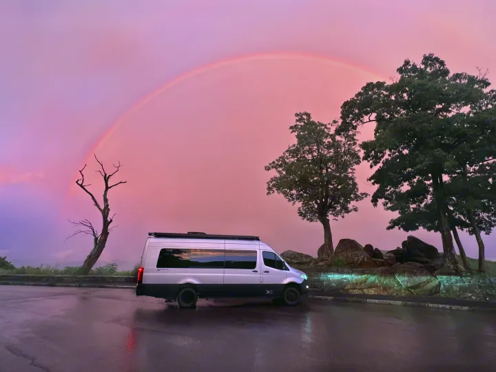 Stunning pink sunset and faint rainbow over an RV rental parked on a wet road, with dark trees and misty mountains.. Airstream Interstate 2023
