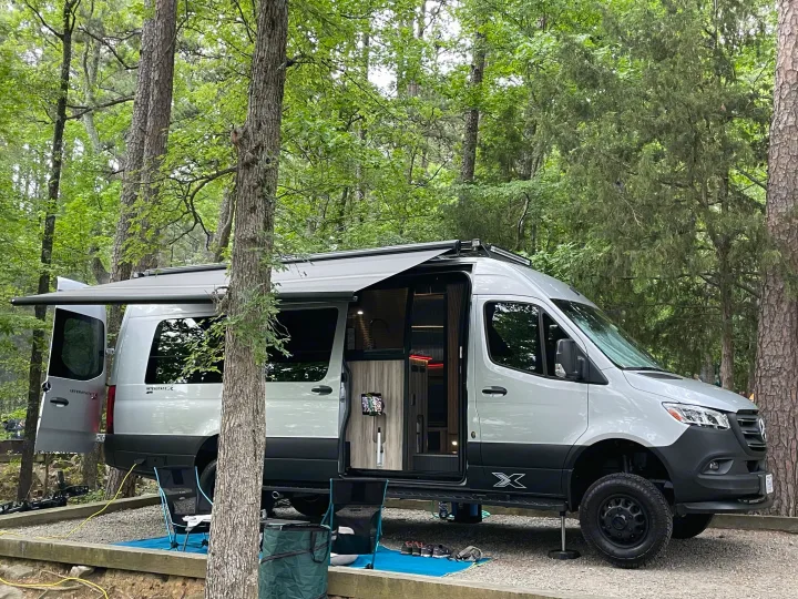This Mercedes-Benz Sprinter camper van, possibly an RV rental, is set up at a wooded campsite with its awning extended and doors open, ready for outdoor living.. Airstream Interstate 2023