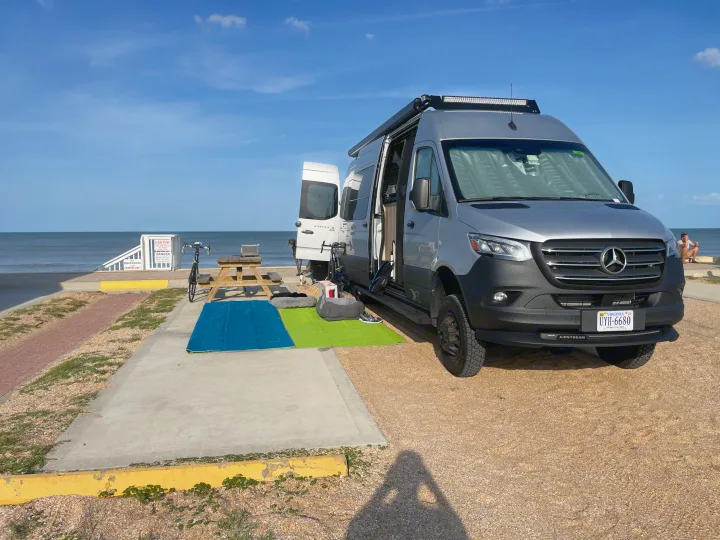 Beachfront RV rental setup: An Airstream van parked, with a picnic table, mats, and dog beds out in Virginia.. Airstream Interstate 2023