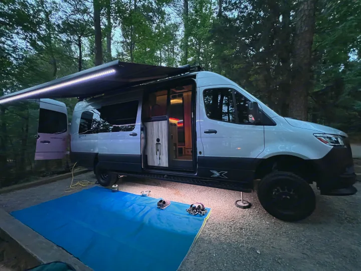 White RV rental camper van set up at a forest campsite at dusk. Awning lights illuminate the outdoor living space.. Airstream Interstate 2023