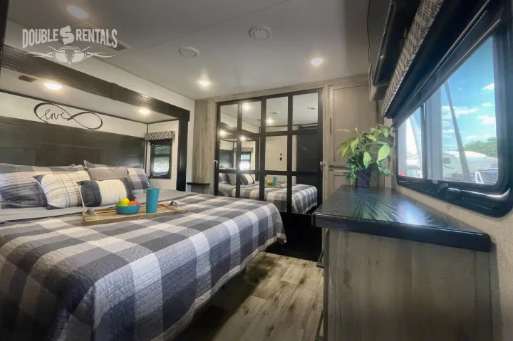 Cozy RV rental bedroom featuring plaid bedding, decorative pillows, a mirrored closet, plant, and a window view.. KZ Durango Gold 2022
