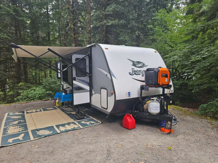 This rv rental camper is set up at a serene forested campsite with its awning, outdoor rug, and generator.. Jayco X213 2016
