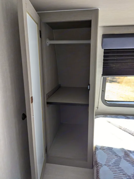 Inside this RV rental, a compact wardrobe with a hanging rod and shelf offers storage next to the bed and window.. Coachmen Apex Nano 2024