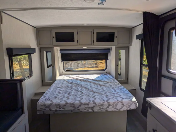 RV rental interior: a bright bedroom with a patterned bedspread, overhead cabinets, and windows showcasing nature outside.. Coachmen Apex Nano 2024