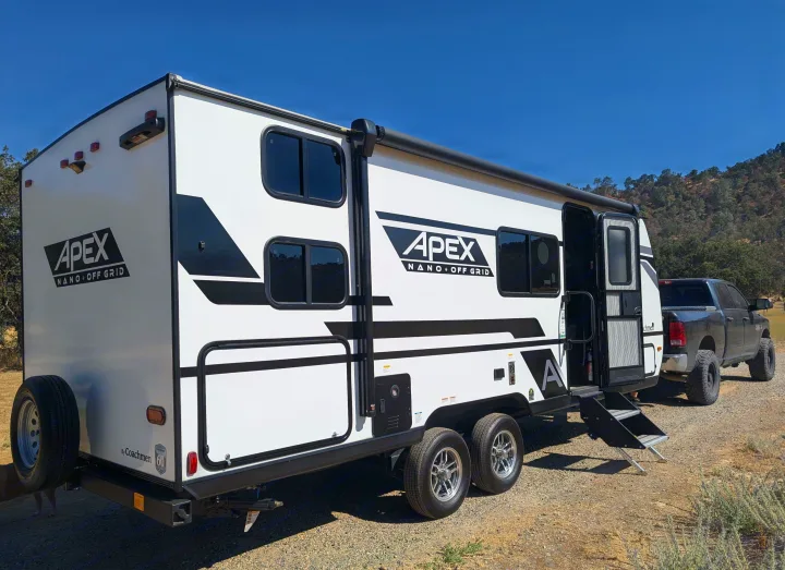 White Apex Nano Off-Grid RV rental with black tow truck on a dirt road, under clear sky, ideal for camping.. Coachmen Apex Nano 2024