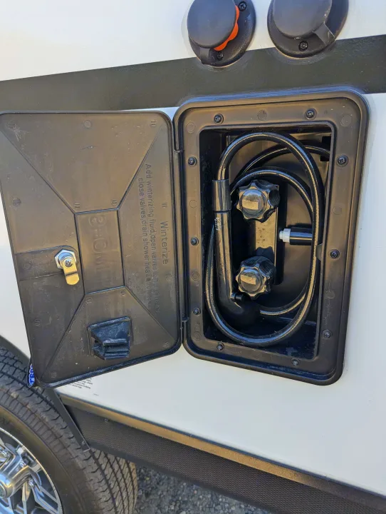 An RV outdoor shower compartment is open, revealing the hose, nozzles, and winterizing instructions for your rv rental adventure.. Coachmen Apex Nano 2024
