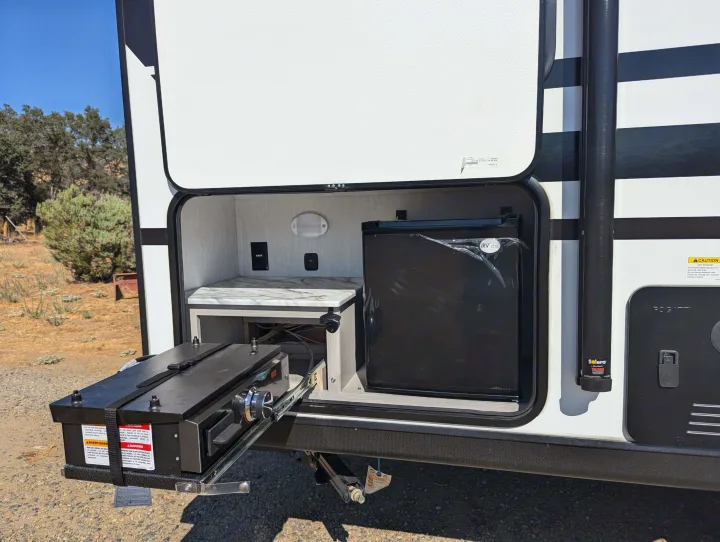 RV rental featuring an outdoor kitchen with a pull-out grill, mini-fridge, and counter area, perfect for campsite cooking.. Coachmen Apex Nano 2024