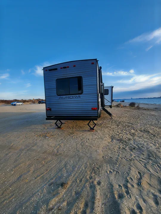 A gray travel trailer RV rental, Aurora model, setup for beach camping. Stabilized on sand near water under a blue sky.. Forest River Aurora Aurora 2024
