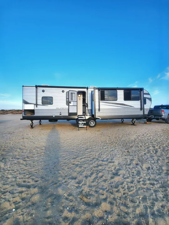 This large RV rental travel trailer, door open, is set up on sand under a clear blue sky, ideal for camping.. Forest River Aurora Aurora 2024