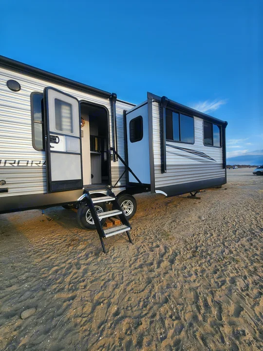 Experience an RV rental! This travel trailer is parked on a sandy spot with its door open, stairs extended, and a slide-out visible.. Forest River Aurora Aurora 2024