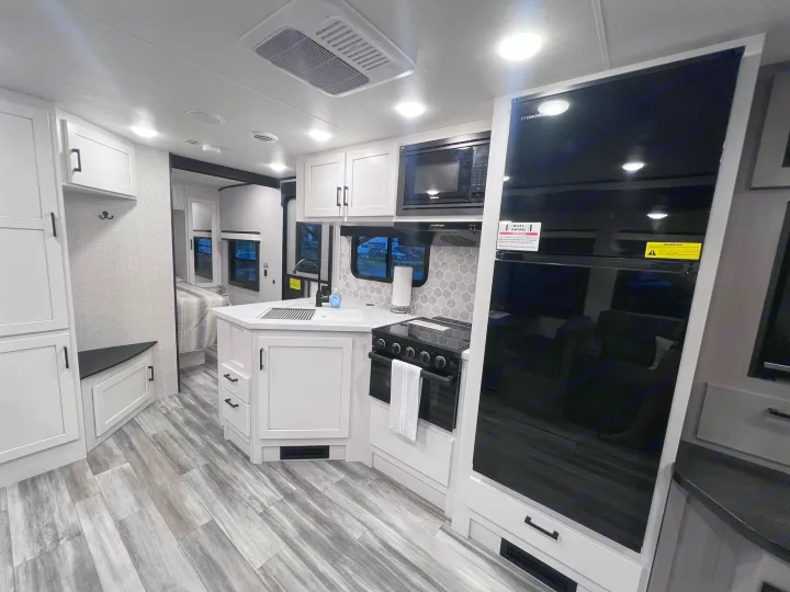 Bright RV rental kitchen with white cabinets, gray wood-look flooring, and major appliances: oven, microwave, large fridge.. Jayco Jay Feather 2024