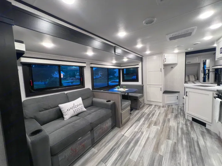 Bright, modern RV rental interior. Features a comfy gray sofa, dinette, and kitchen space with wood-look flooring.. Jayco Jay Feather 2024