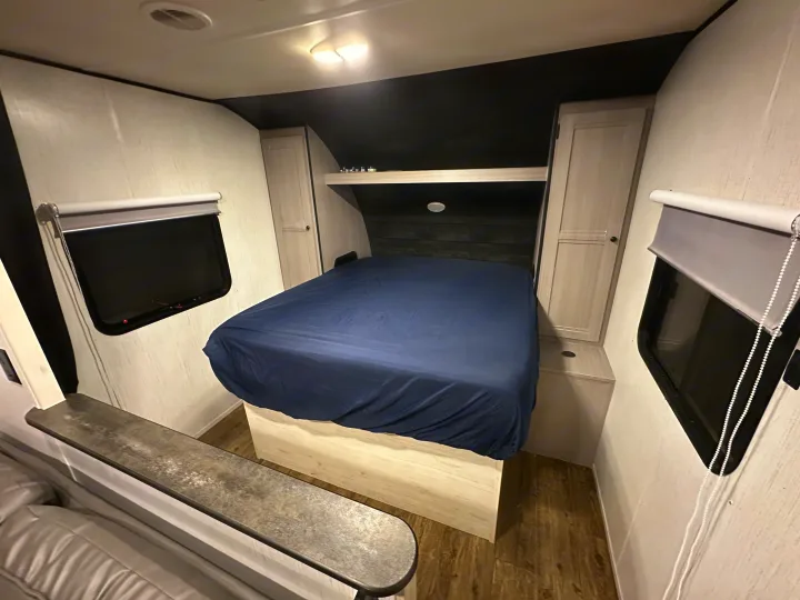 Modern RV rental bedroom featuring a bed with a blue cover, side storage, windows with shades, and light wood-look flooring.. Heartland RVs Trail Runner 2022
