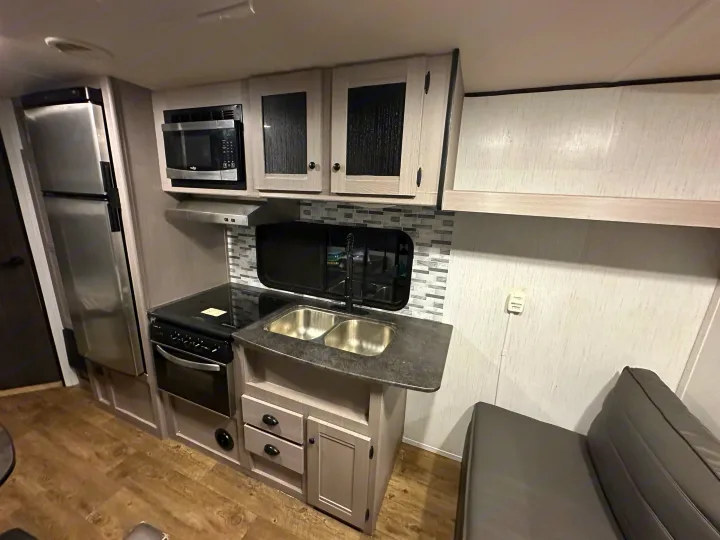 Modern RV rental kitchen with fridge, microwave, oven, double sink, and wood-look floor. Light cabinets & gray backsplash.. Heartland RVs Trail Runner 2022