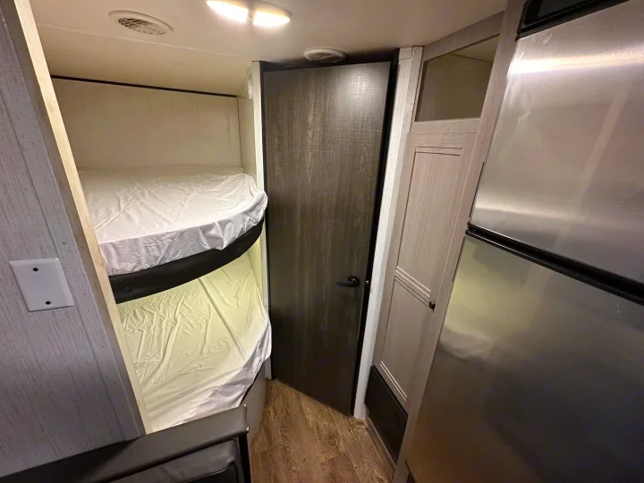 RV rental interior: cozy bunk beds, a dark door, light wood cabinets, and a shiny stainless steel refrigerator unit.. Heartland RVs Trail Runner 2022