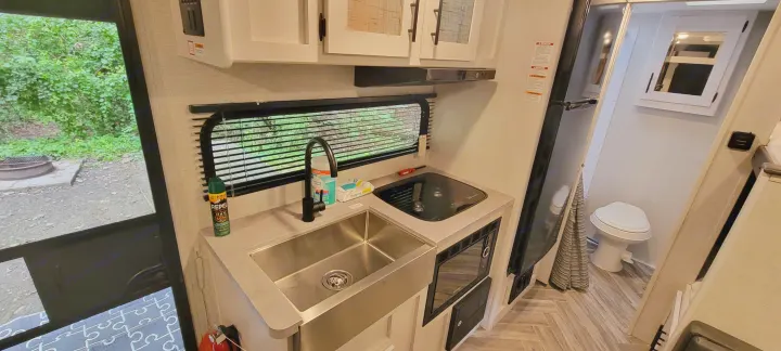 View inside an RV rental: a compact kitchen with sink, stove, microwave, fridge, bathroom, and campsite view.. Dutchmen Coleman Rubicon 2021