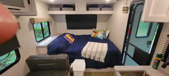 Cozy RV rental interior: A compact bedroom featuring a navy bed, pillows, windows with nature views, and overhead storage.. Dutchmen Coleman Rubicon 2021