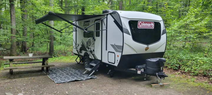 A Coleman travel trailer **RV rental** parked at a serene wooded campsite, featuring an extended awning and picnic area.. Dutchmen Coleman Rubicon 2021