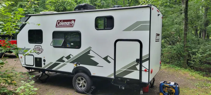 This Coleman RV rental, a compact travel trailer, is set up with a generator at a serene wooded campsite.. Dutchmen Coleman Rubicon 2021