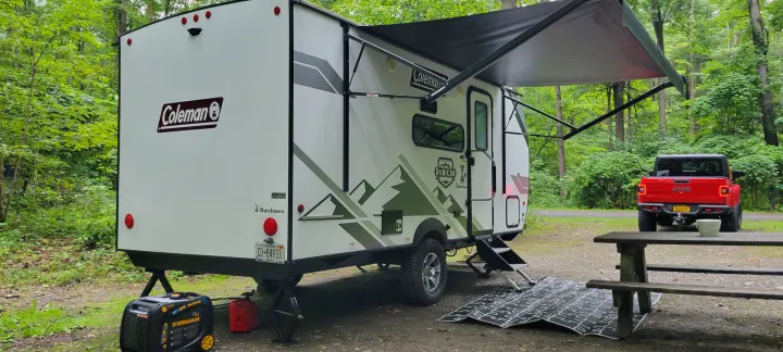 A white Coleman travel trailer RV rental is set up at a wooded campsite, with a red Jeep and generator nearby.. Dutchmen Coleman Rubicon 2021