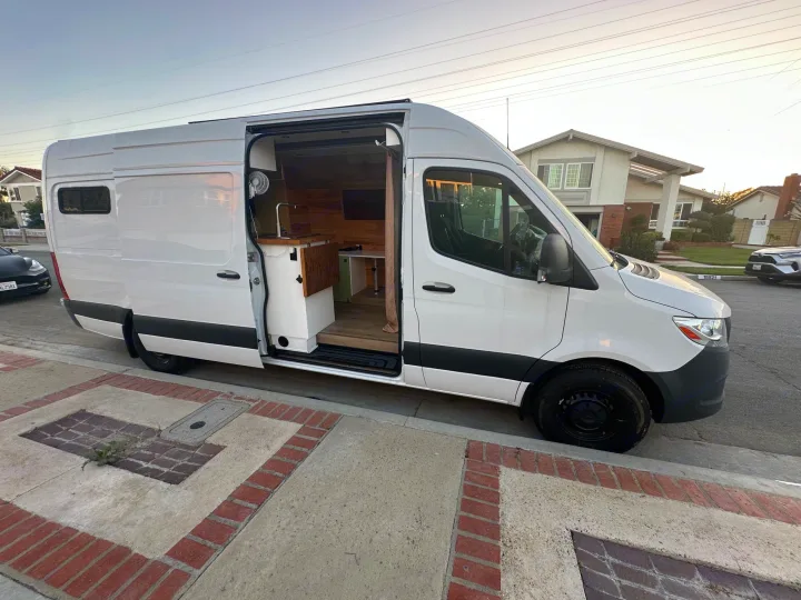 Stylish white camper van conversion with open door revealing a cozy wood interior and kitchen setup, ideal for rv rental.. Mercedes-Benz Sprinter 2022