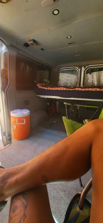 RV rental interior features a bed, orange cooler, and camping chairs, viewed from a person's seated perspective.. Dodge Sprinter Van 2007