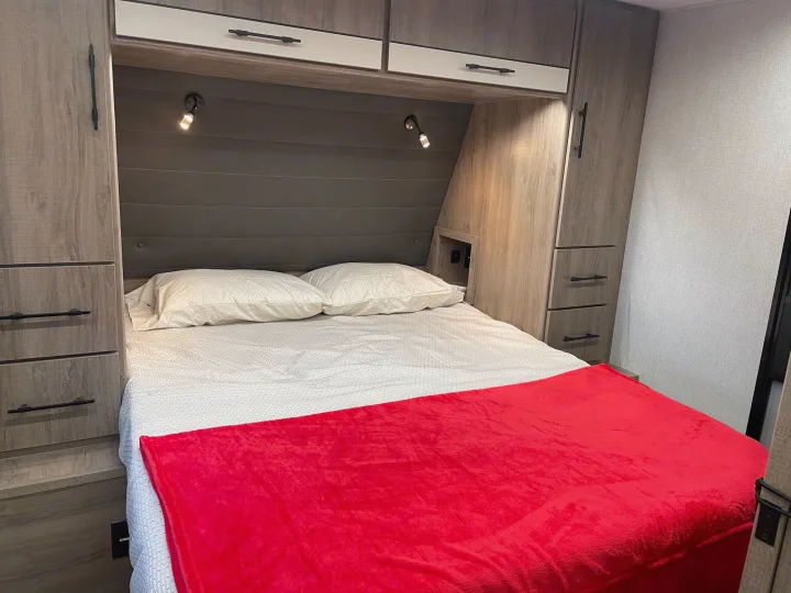 Comfortable RV rental bedroom featuring built-in storage, reading lights, and a cozy red blanket for travel.. Grand Design Imagine XLS 2024