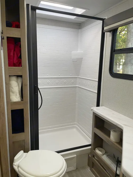 This modern RV rental bathroom boasts a clean white shower with a black frame, toilet, and built-in storage.. Grand Design Imagine XLS 2024