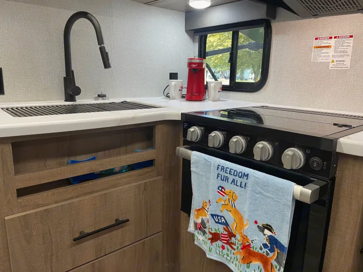 Modern RV rental kitchen featuring a sink, stove, coffee maker, and "Freedom Fur All!" towel. Compact and functional.. Grand Design Imagine XLS 2024