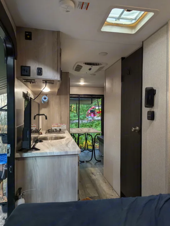 Inside an RV rental, a compact kitchen with sink, stove, and TV. A dinette and large rear window show lush forest views.. KZ Escape Hatch 2022
