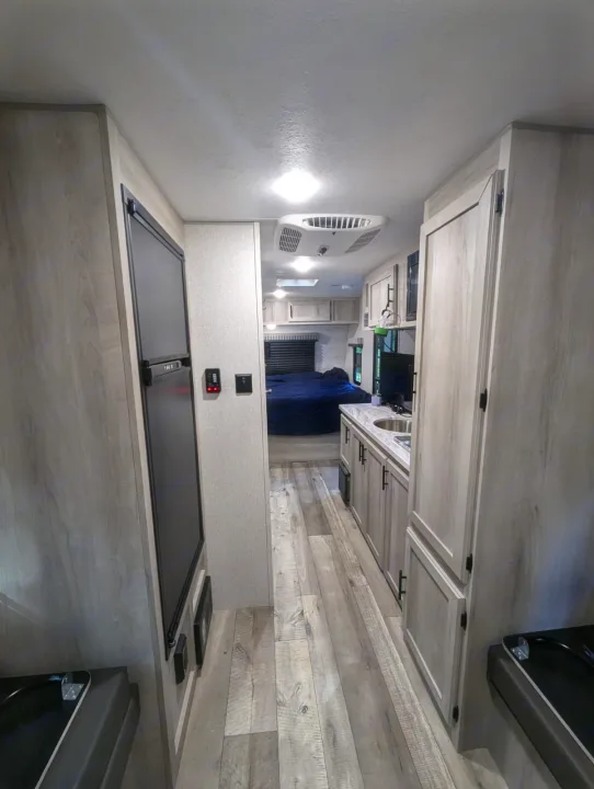Modern RV rental interior: hallway leads to a galley kitchen, refrigerator, and rear bedroom area with dark bed.. KZ Escape Hatch 2022