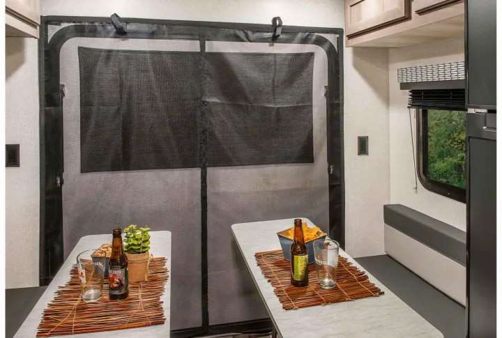 RV rental interior with a large black mesh screen, two tables holding drinks/snacks, bench seating, and window with trees.. KZ Escape Hatch 2022
