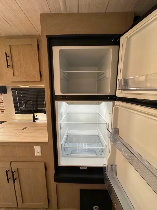 Inside an rv rental, an open, empty fridge/freezer unit stands next to a light-wood kitchen counter, sink, and cabinets.. Forest River Wildwood Platinum 2024