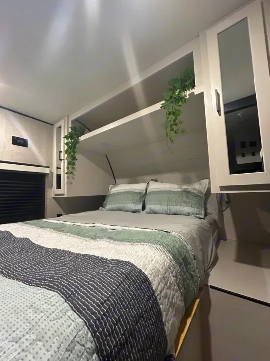 Cozy modern RV rental bedroom featuring a bed with striped bedding, overhead shelves with hanging plants, and white cabinetry.. Jayco Jay Flight 2024