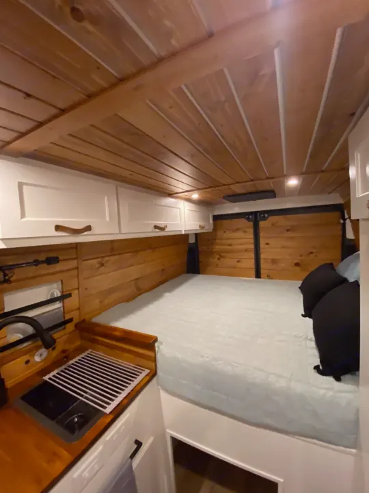 Well-appointed RV rental camper van interior. Shows a comfortable bed, wood-paneled walls/ceiling, white cabinets, and a kitchen sink.. RAM Promaster 2300 2023