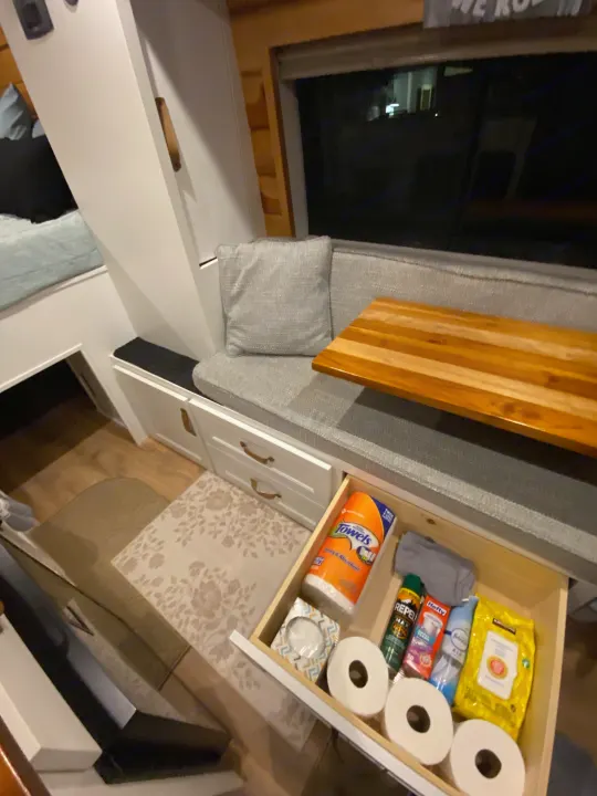 RV rental interior showcases clever storage: seating, a bed, and an open drawer stocked with essentials like paper towels and toilet paper.. RAM Promaster 2300 2023