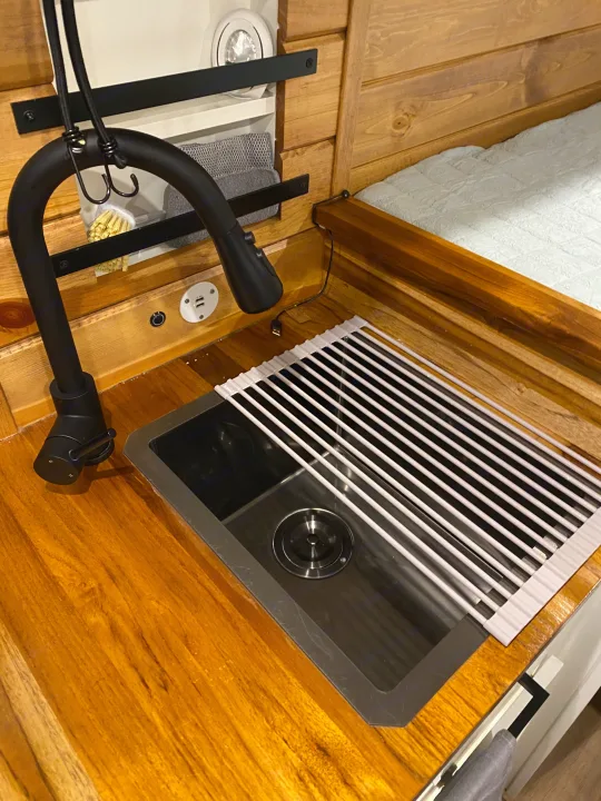 RV rental interior: cozy kitchen with wooden counter, sink, black faucet & drying rack. USB port and bed nearby.. RAM Promaster 2300 2023