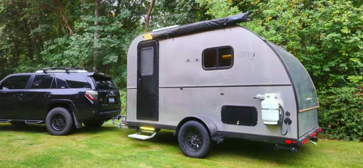 Black SUV hitched to a silver teardrop RV camper, ready for adventure. Explore nature with an RV rental!. Bend Teardrop Tall Boy 2023