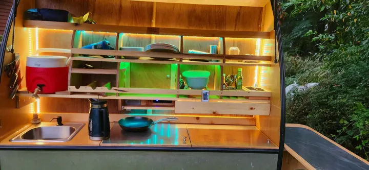 Bright, organized camper kitchen setup, ideal for an RV rental. Features shelves, LED lights, sink, cooler, and cooking pan.. Bend Teardrop Tall Boy 2023