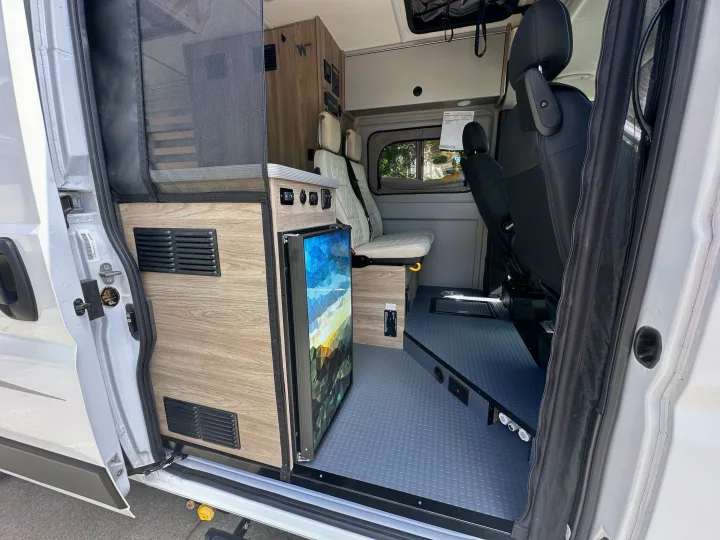 Compact RV rental interior featuring a fridge, adaptable seating, and wood finishes, ready for your next adventure.. Winnebago Solis 2025