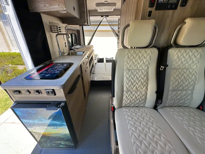 Modern rv rental interior: compact kitchen with sink, stovetop & fridge; comfortable passenger seats. Rear door open to outside.. Winnebago Solis 2025
