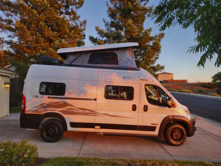 A white Winnebago Solis camper van with a raised pop-top roof, bathed in golden sunset light on a driveway, ideal for an RV rental.. Winnebago Solis 2025