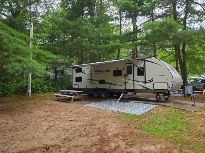 A large Coachmen RV rental is set up at a shaded forest campsite with a picnic table and outdoor mat.. Coachmen Freedom Express Liberty Edition 2016